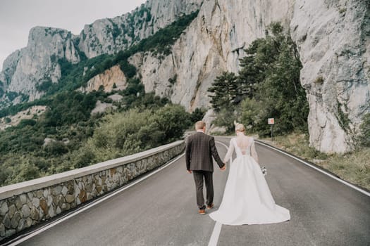 A bride and groom are standing on a road, holding hands. The bride is wearing a white dress and the groom is wearing a suit. The scene is set in a mountainous area, with a rocky road in the background