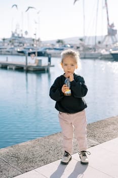 Little girl drinks juice through a straw from a bottle while standing on the boardwalk. High quality photo