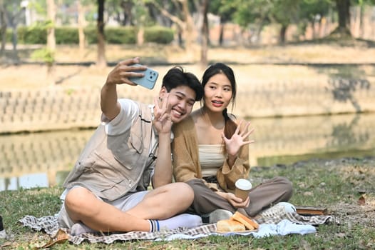 Shot of a cheerful young couple sitting on on blanket in park and talking selfie with smartphone.