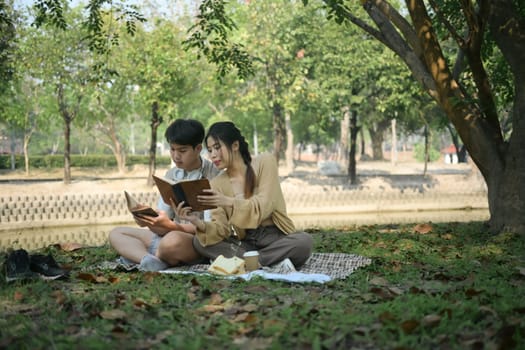 Relaxed young couple spending time together outdoor reading book on picnic blanket in the park.