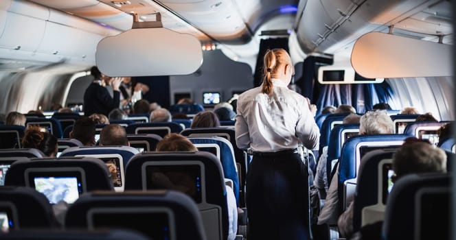 Interior of airplane with passengers on seats and stewardess in uniform walking the aisle, serving people. Commercial economy flight service concept