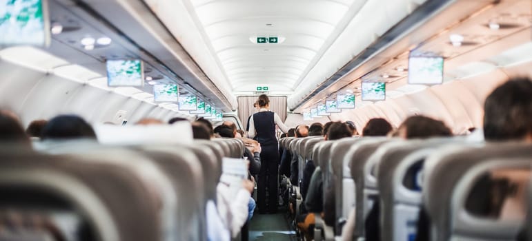 Interior of airplane with passengers on seats and stewardess in uniform walking the aisle, serving people. Commercial economy flight service concept