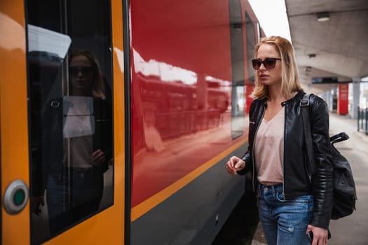 Young blond woman in jeans, shirt and leather jacket wearing bag and sunglass, embarking red modern speed train on train station platform. Travel and transportation