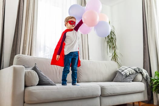 Young child in superhero costume with red cape and eyemask holding balloons on a couch. Imagination and play concept. Design for childrens party invitations and celebration posters with copy space.
