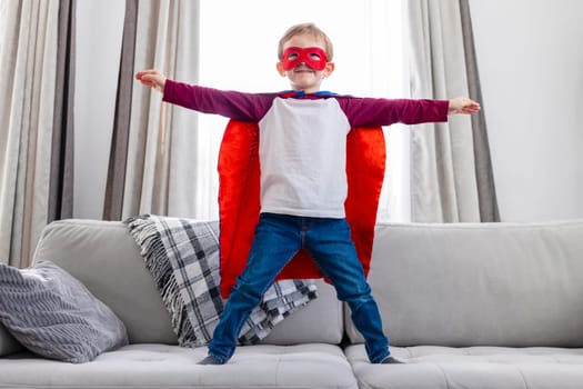Child in superhero outfit standing confidently on sofa. Home interior setting with natural light. Creativity and childhood play concept. Design for greeting card, invitation, poster.