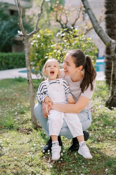 Little laughing girl sitting on smiling mother lap squatting in garden. High quality photo