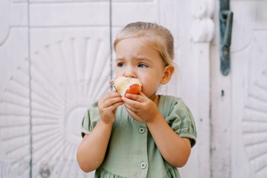 Little girl gnaws a red apple while standing near a white carved wooden door and looks away. High quality photo