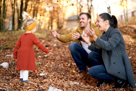 Happy mom and dad sprinkle dry leaves on a little girl sitting in the autumn forest. High quality photo
