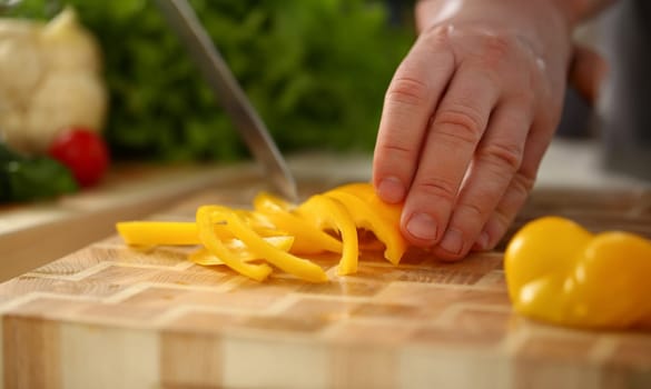 Cook holds knife in hand and cuts on cutting board yellow pepper for salad or fresh vegetable soup with vitamins. Raw food and vegetarian recipe book in modern society popular concept.