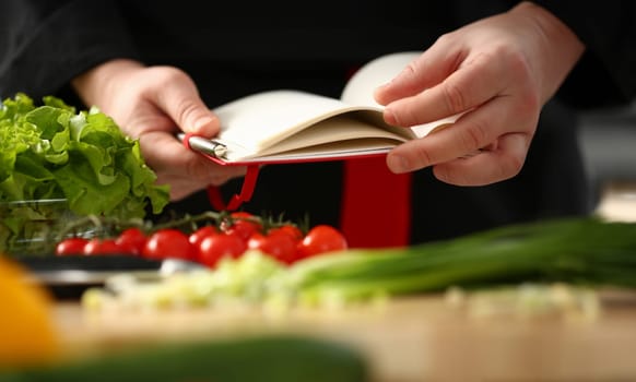 Male cook hand hold siver pen and wright open book of recipes on background of vegetables. Chef writes new combination of taste in home magazine shopping organizer concept list