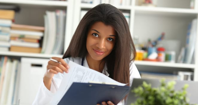 Beautiful black smiling female doctor hold clipboard pad and fill something with silver pen. Physical illness prevention prescribe remedy ward round therapeutist assistance healthy lifestyle concept