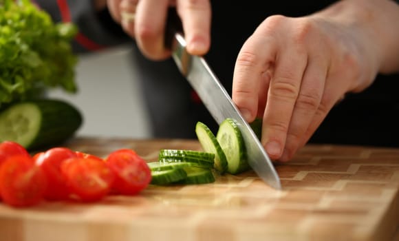 Cook holds knife in hand and cuts on cutting board green cucumber for salad or fresh vegetable soup with vitamins. Raw food and vegetarian recipe book in modern society popular concept.