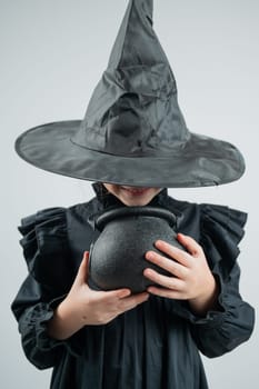 Portrait of a little Caucasian girl in a witch costume holding a cauldron on a white background. Vertical photo
