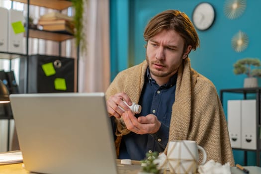 Sick ill young businessman suffering from cold flu or allergy working on laptop at home office. Manager freelancer man wrapped in blanket blows sneezes wipes snot into napkin. Coronavirus quarantine