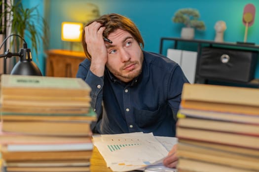 Exhausted dissatisfied Caucasian businessman moving stacks of books and reviewing documents reports working hard in home office. Stressed tired bored freelancer touching head in despair. Overload