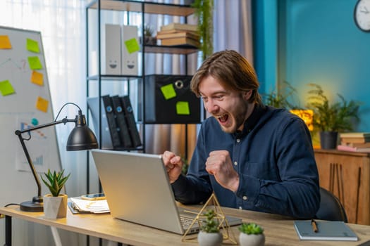 Young Caucasian businessman working on laptop netbook shocked by sudden victory, winning, goal achievement, good career news. Excited freelancer guy clenching fists at home office.
