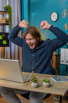 Young happy businessman working on laptop netbook shocked by sudden victory, winning, goal achievement, good career news. Excited amazed freelancer guy clenching fists at home office. Vertical