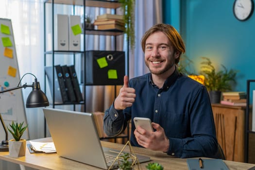Smiling businessman using smartphone app online pointing showing like thumbs up remote distance job working from home office. Man freelancer guy e-learning texting messages content on social media.