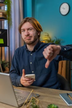 Dislike. Upset sad Caucasian businessman using smartphone at home office showing thumbs down sign gesture, expressing discontent, disapproval dissatisfied bad work. Displeased guy freelancer.