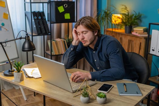 Bored sleepy businessman worker working on laptop computer leaning on hand sleeps at home office desk. Exhausted tired Caucasian young man freelancer workaholic guy. Employment, occupation, workless.