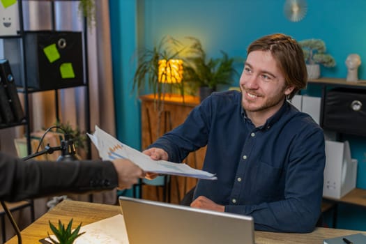Caucasian freelancer businessman receiving documents from boss and working with financial graph, calculate taxes for charts and reports in home office. Guy working on laptop, planning expenses budget.