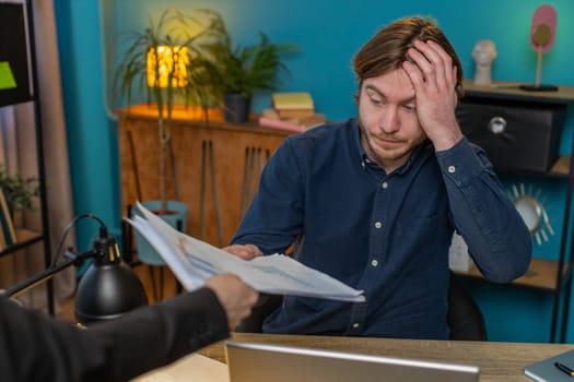 Tired exhausted Caucasian freelancer businessman receiving documents from boss experiences difficulties due to fatigue sitting at table in home office. Stressed guy looking frustrated using laptop