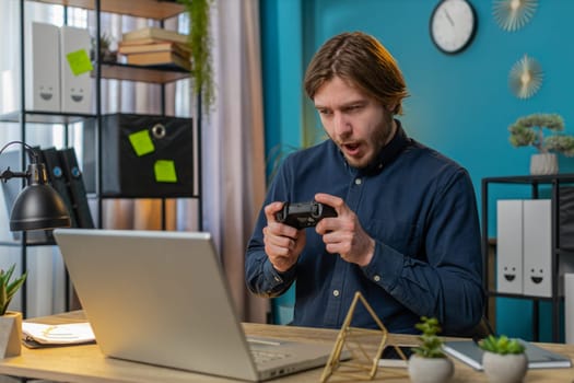 Excited Caucasian businessman celebrating victory in video game on laptop spending leisure time at home office. Happy freelancer guy playing video games holding controller. Relaxing taking a break