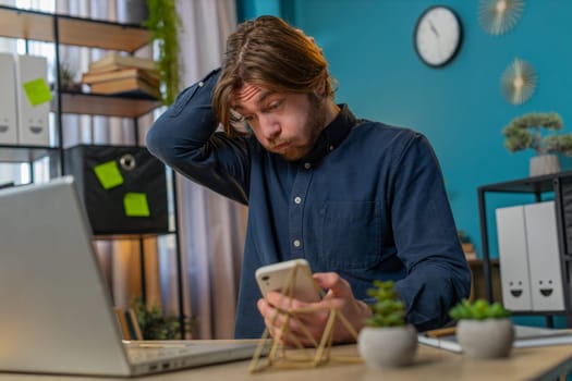Amazed young businessman using smartphone typing browsing, loses becoming surprised sudden lottery results, bad news, fortune loss fail at home office desk workplace. Bearded freelancer worker man.