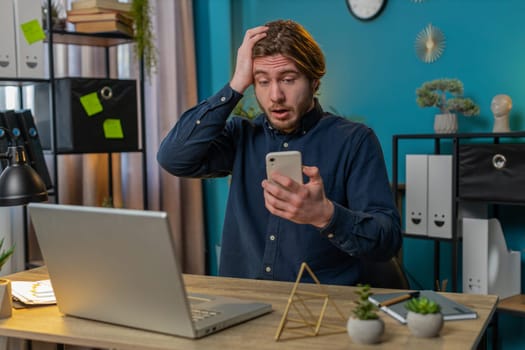 Caucasian young businessman using smartphone typing browsing, loses becoming surprised sudden lottery results, bad news, fortune loss fail at home office desk workplace. Bearded freelancer worker man