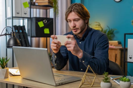 Young freelancer relaxing during break and playing online video games on smartphone during work day at home. Man enthusiastically playing racing or shooter video games on smartphone sitting at table.