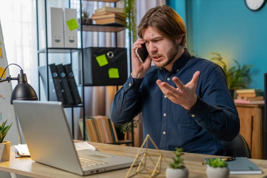 Angry businessman freelancer answering to client on mobile phone call, using smartphone and laptop. Conversation. Caucasian man guy working online remote distance job, talking at home office desk.