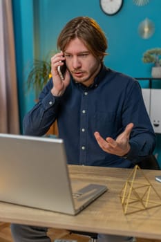 Angry businessman freelancer answering to client on mobile phone call, using smartphone and laptop. Conversation. Caucasian man guy working online remote distance job, talking at home office desk.