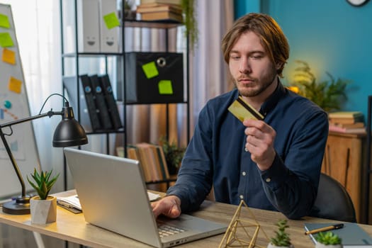 Young Caucasian businessman using bank credit card and laptop for online shopping payments at home office. Business transaction. Strict guy freelancer purchases with netbook E-banking app service.