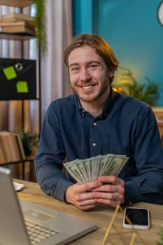 Young businessman guy holding fan of cash money dollar banknotes showing thumbs up and pointing at laptop, celebrate, success career, online income, wealth at home office table. Remote distant working