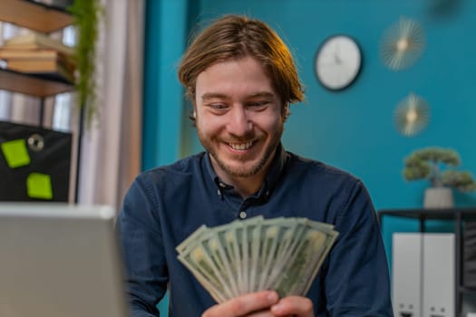 Amazed businessman guy holding fan of cash money dollar banknotes showing thumbs up and pointing at laptop, celebrate, success career, online income, wealth at home office. Remote distant working.