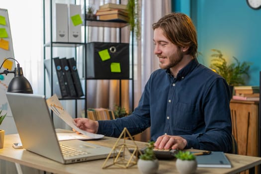 Focused young office worker working with schedules and financial graphics while using calculator and laptop sitting at home office desk. Happy businessman freelancer compare and analyze graphs charts.