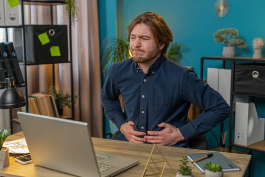 Disappointed Caucasian freelancer feeling overeating during work process on laptop in home office desk. Upset businessman freelancer guy man touching stomach and experiencing discomfort and sharp pain
