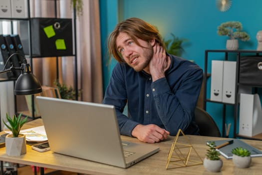 Tired Caucasian businessman suffers from spine neck muscle pain at home office workplace working on laptop netbook. Overworking. Bearded freelancer sick man guy attempts to relieve pain by massaging