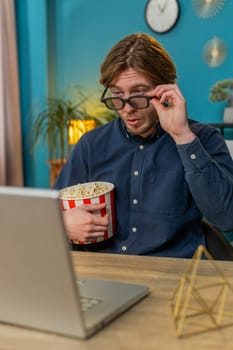 Businessman freelancer taking break from work wearing 3D glasses eating popcorn and watching movie on laptop. Laughing young man guy sitting at home office desk having snacks during break. Vertical