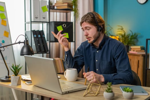Caucasian businessman wearing headset, freelance worker, call center or support service operator helpline, talking with client or colleague communication support. Happy man sits at home office desk.