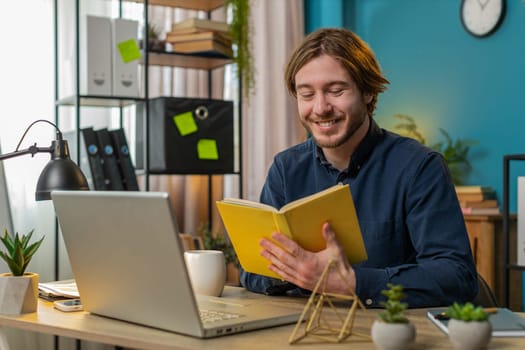Caucasian young businessman reading interesting book, turning pages smiling enjoying literature, taking a rest after work. Happy cheerful freelancer guy with laptop relaxing at home office table.
