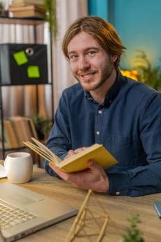 Young businessman reading interesting book, turning pages smiling enjoying literature, taking a rest after work. Happy cheerful freelancer guy with laptop relaxing at home office table. Vertical