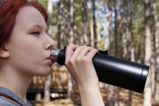 girl drinks tea from a black thermos in the forest, close-up side view.