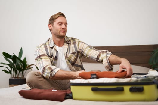 Portrait of handsome young man, tourist unpacking clothes in hotel room, staying in hostel on vacation, travelling, sitting with luggage on bed.