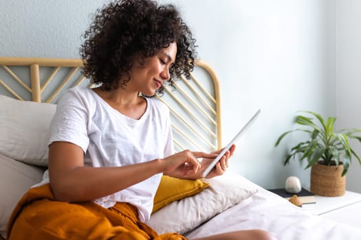 Young African American woman using tablet lying down relaxing on bed at home cozy bedroom. Technology concept.