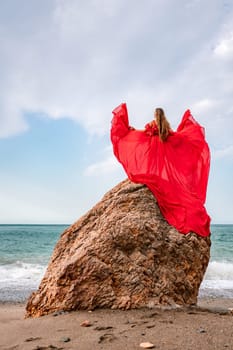 woman sea red dress. Woman with long hair on a sunny seashore in a red flowing dress, back view, silk fabric waving in the wind. Against the backdrop of the blue sky and mountains on the seashore