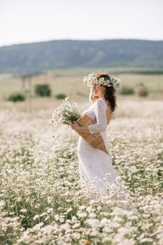 Happy woman in a field of daisies with a wreath of wildflowers on her head. woman in a white dress in a field of white flowers. Charming woman with a bouquet of daisies, tender summer photo.