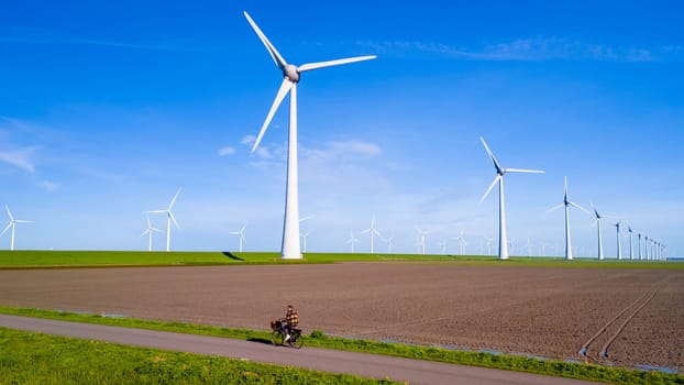 A man rides an electric bike bicycle down a dusty road alongside a sprawling wind farm with rows of turbines, set against a clear blue sky in the Netherlands Flevoland during spring.