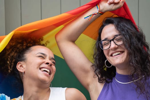 Headshot of lesbian young couple with positive mood laughing and smiling with LGBTQ rainbow flag.