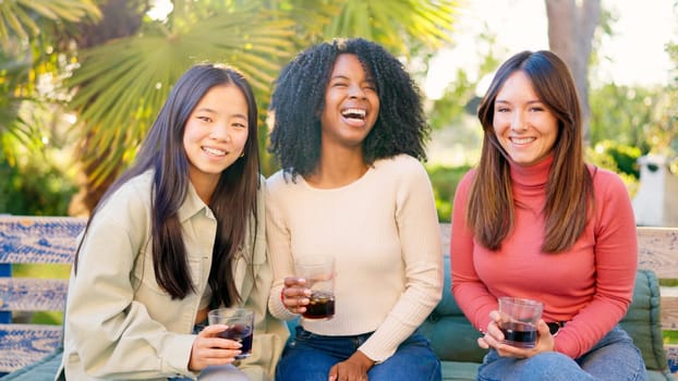 Three multi-ethnic friends smiling at camera while drinking wine in the garden during sunset